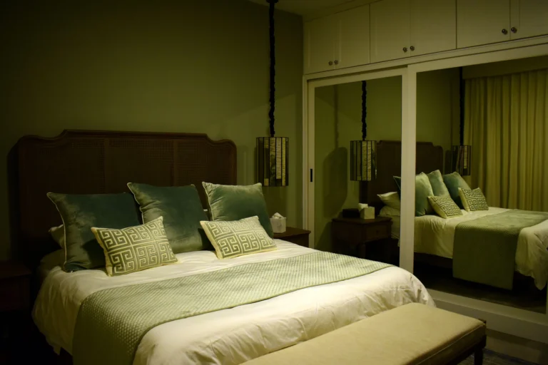 A dim, warmly lit bedroom at night with a made bed, green velvet cushions, mirrored wardrobes and curtains — small bedroom sleep ideas that bring deeper rest