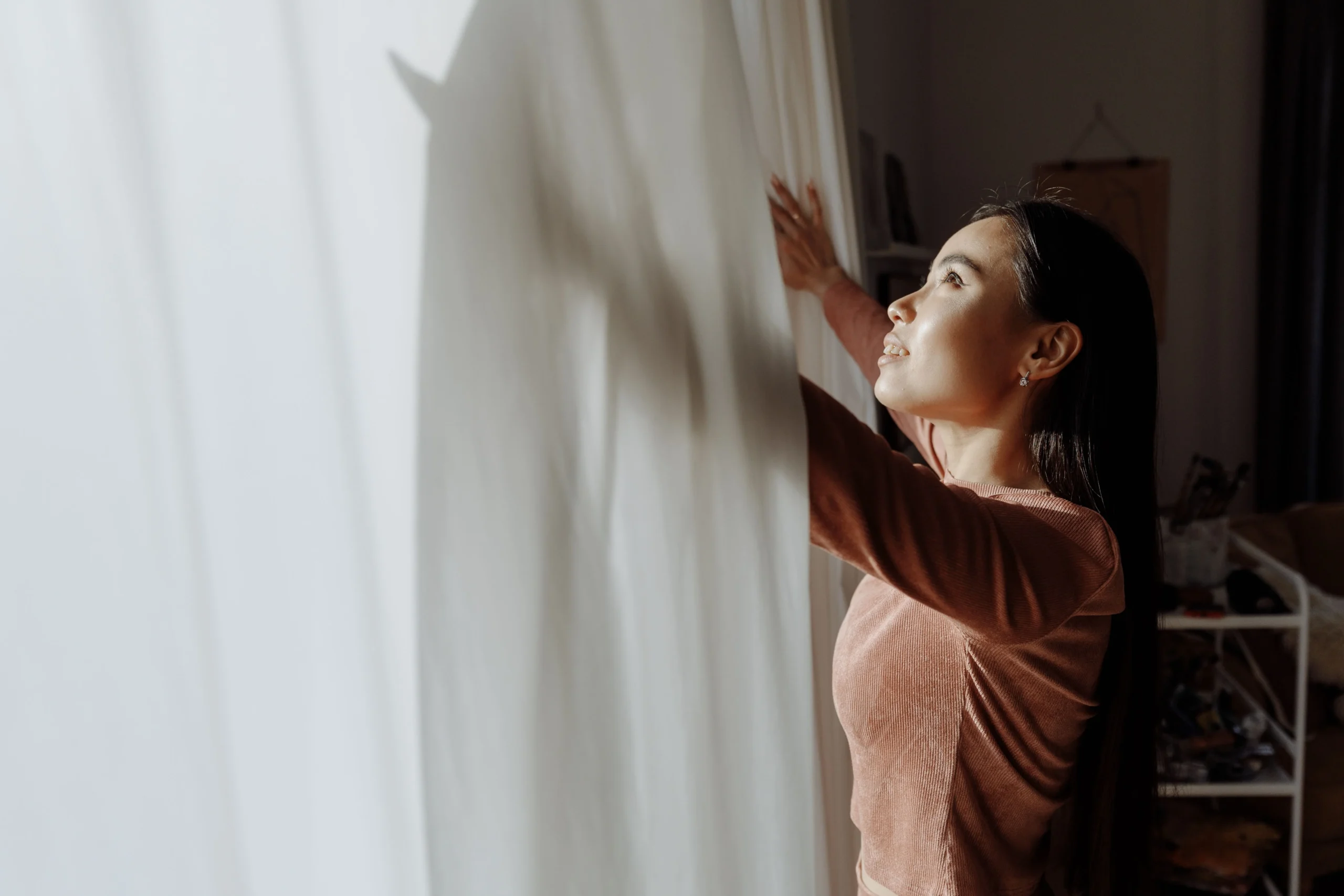 A woman opening a white curtain, embracing daylight indoors — 9. morning light first, before the phone