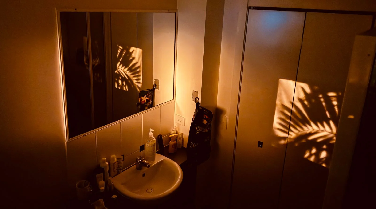 Warm small bathroom in soft morning light with palm-leaf shadows on the wall, mirror and sink — small bathroom morning ideas for a calm get-ready routine