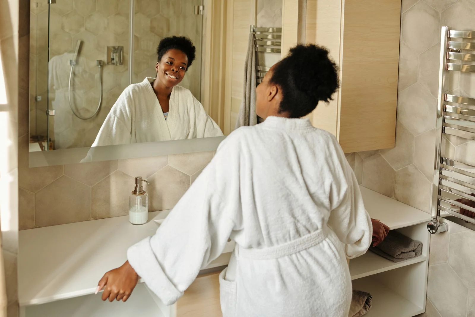 Woman in a bathrobe smiling at her reflection in a modern bathroom mirror — mirror and light first in a small bathroom morning routine