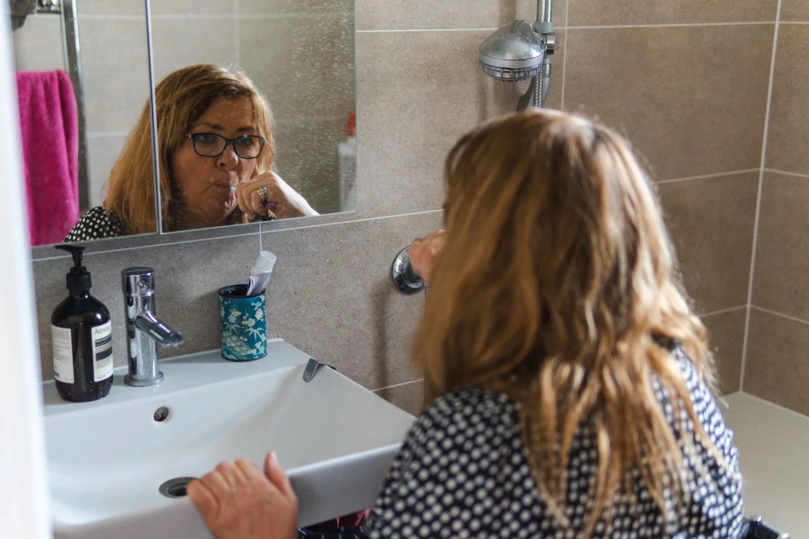 Woman brushing her teeth reflected in a bathroom mirror — same-order get-ready routine in a small bathroom