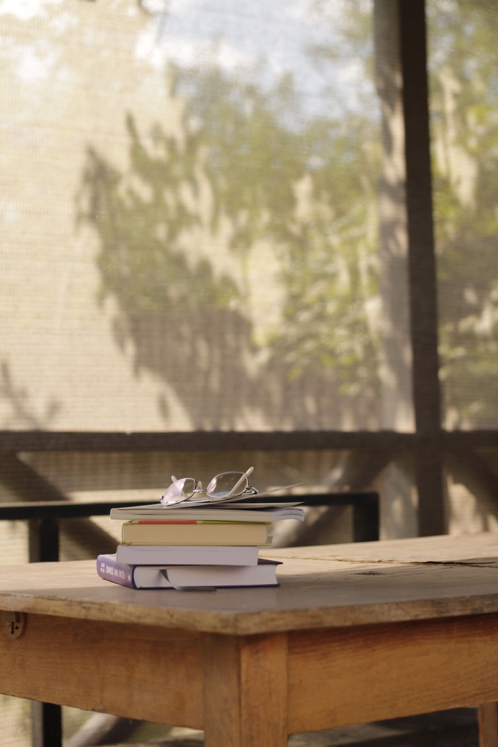 Tidy stack of books with reading glasses on a wooden table by a sunny window