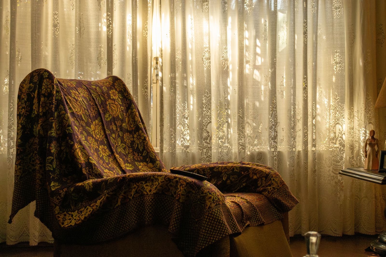 Vintage armchair draped with a floral throw beside a sunlit lace-curtained window