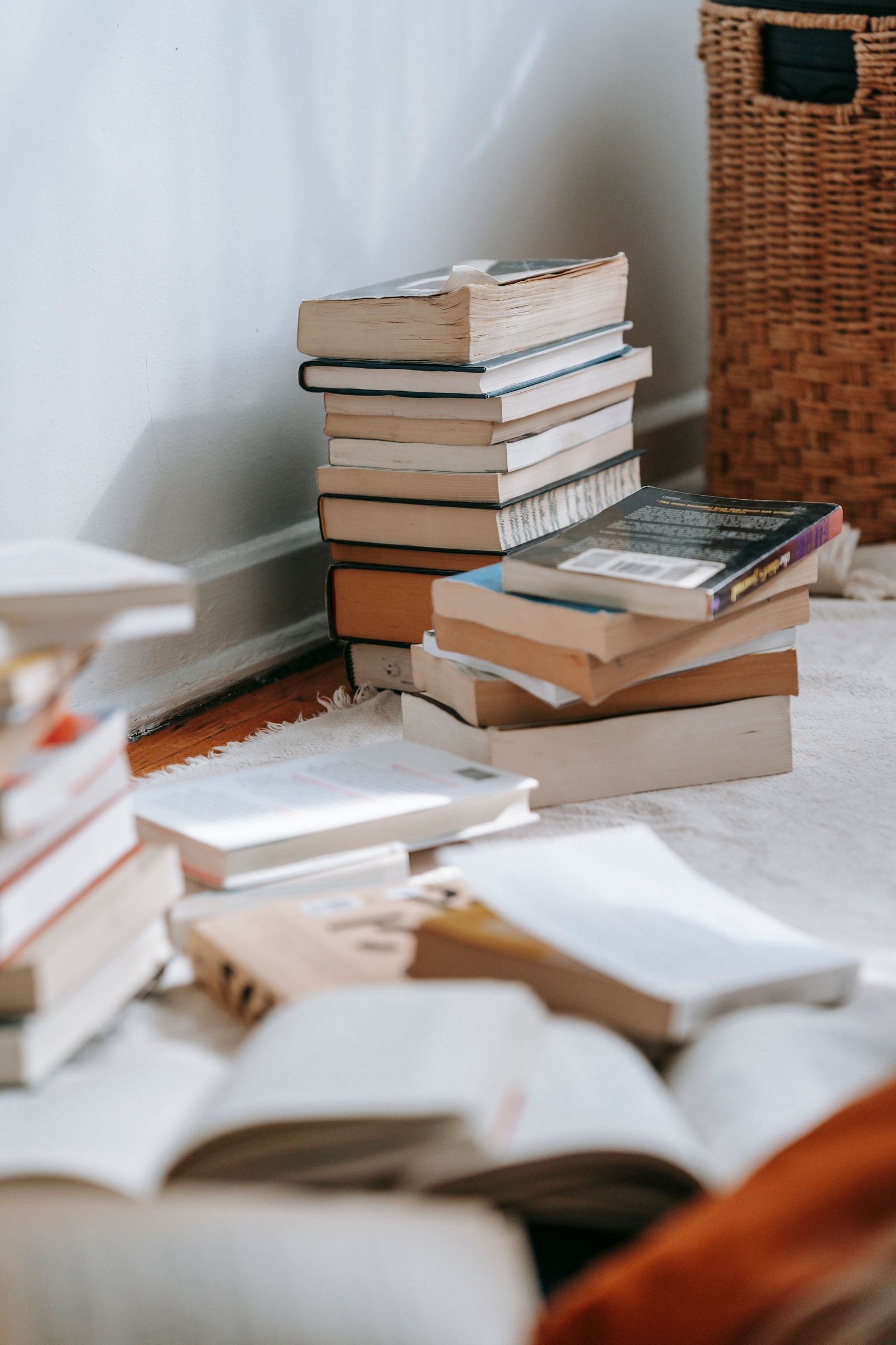 Stacks of books on a white rug with a wicker basket beside them in a bright corner