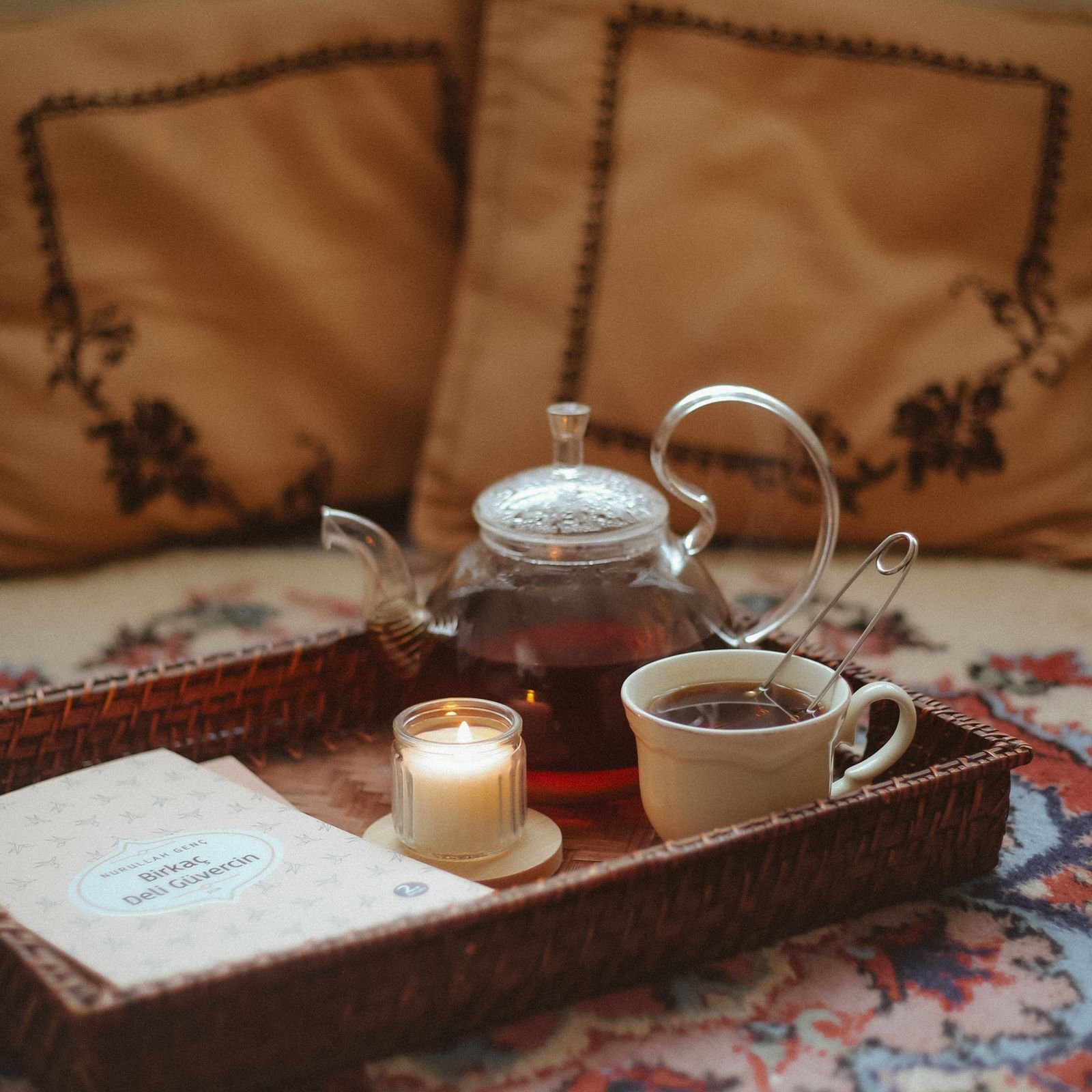 Wicker tray with glass teapot, cup of tea, lit candle, and a book