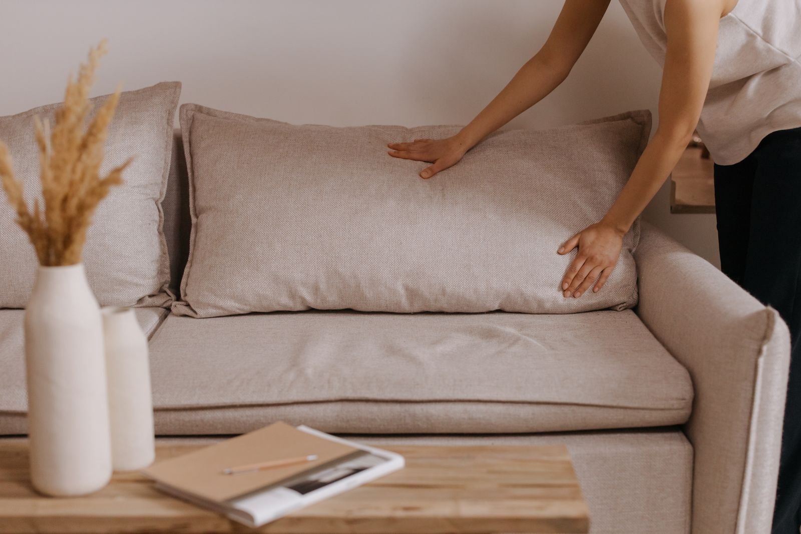 Person straightening a pillow on a beige sofa in a tidy minimalist living room