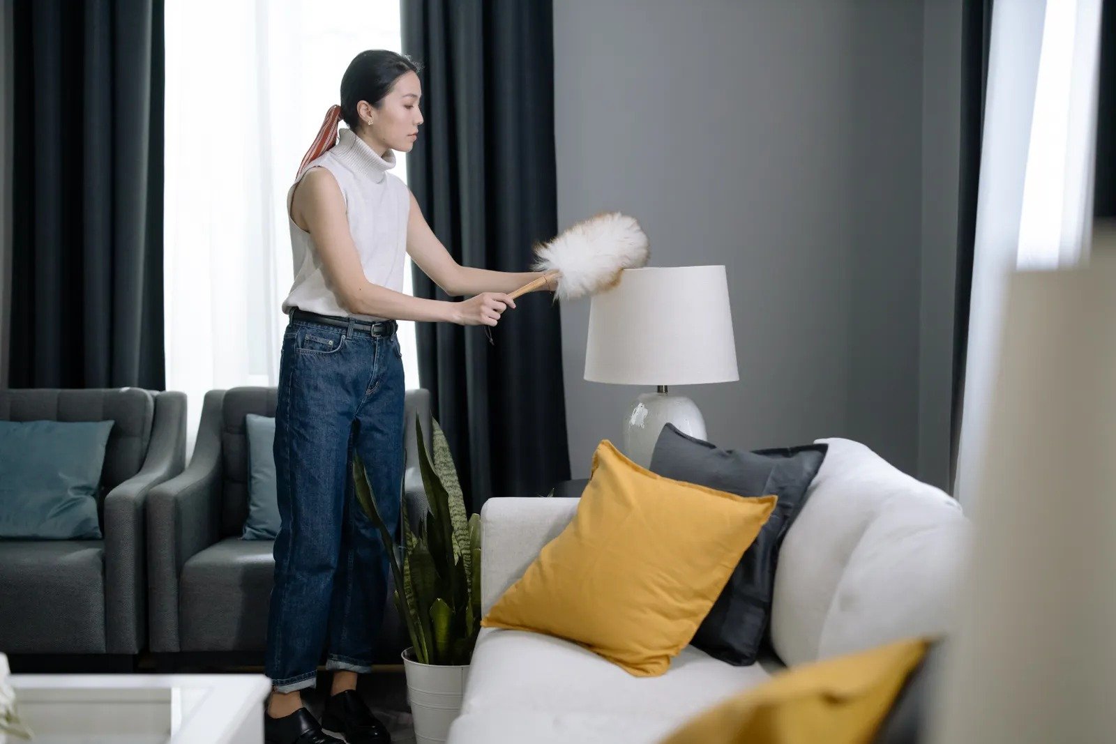 Woman dusting a lampshade with a feather duster in a tidy modern living room